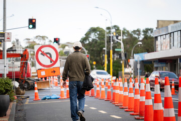 Man walking on the road with orange traffic cones diverting the traffic. Bus stop blocked by the traffic cones. Roadworks in Auckland. - 792699237