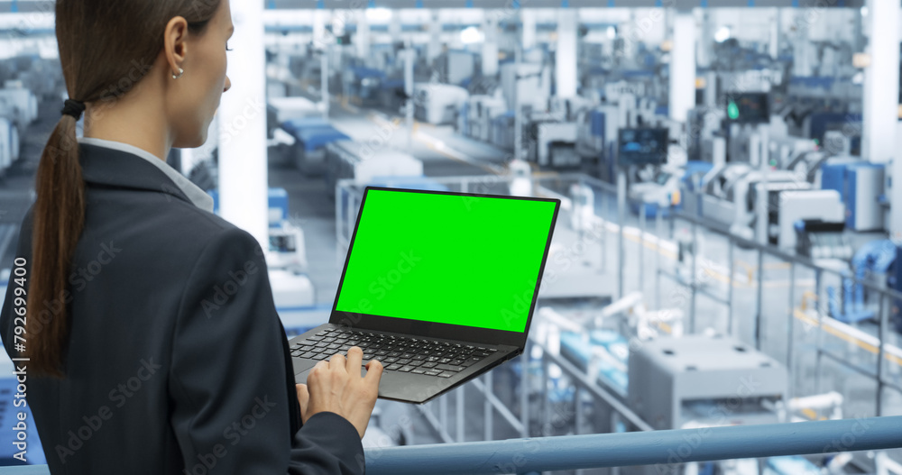 Poster Young Female Engineer Using Laptop Computer with Green Screen Mock Up Display at an Electronics Factory. Automated Manufacturing Machines and AI Robots Are Monitored Online
