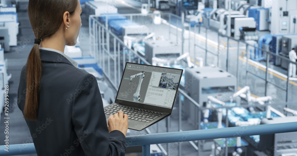 Leinwandbilder Young Female Engineer Standing on a Platform, Using Laptop Computer at an Electronics Factory. Machines are Undergoing Maintenance, Technician Monitoring Updates Through Online Software
