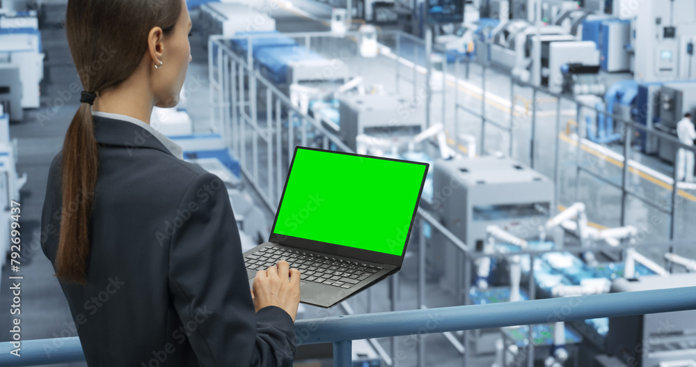 Poster Female Technician Using Laptop Computer with Green Screen Chroma Key Mock Up Display and Looking Around a Factory Facility with Robotic Arms Producing Modern Electronic Components for Tech Industries