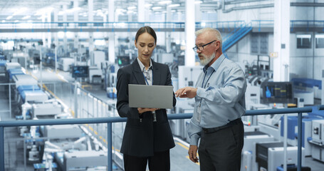 Experienced Male and Female Engineers Standing on a Platform at a Modern Industrial Factory, Using Laptop Computer and Discussing Production of Modern Electronics for Artificial Intelligence Machines