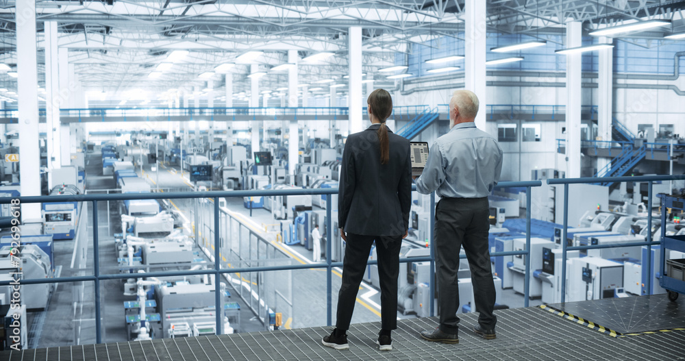 Poster Male Engineer and Female Project Manager Standing on a Platform, Talking, Using Laptop Computer and Monitoring Production at a Modern Automated Electronics Manufacture with AI Robotic Arms