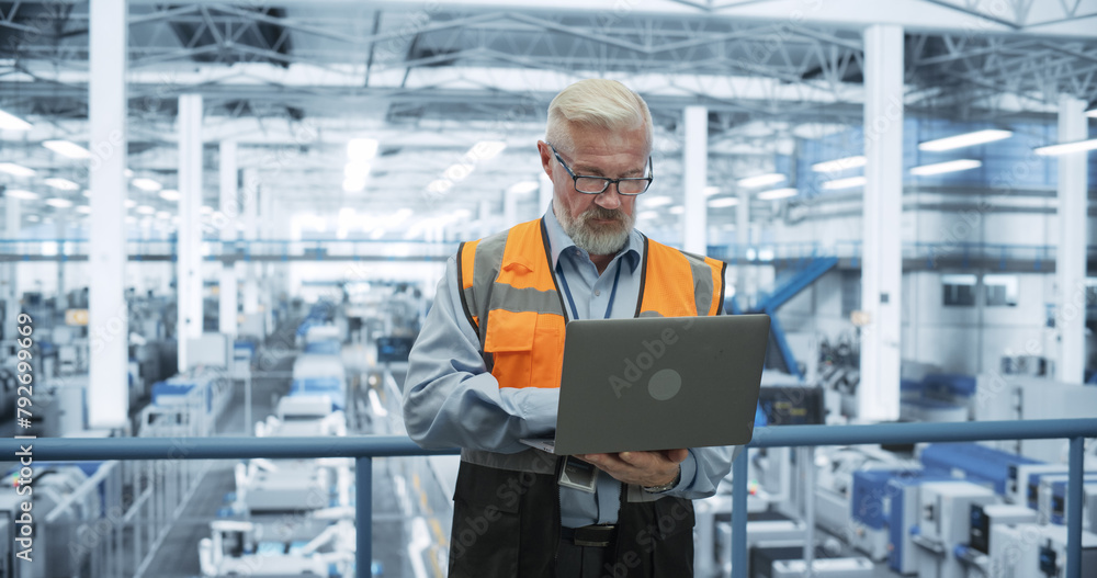 Poster Senior Engineer Standing on a Platform, Using Laptop Computer at an Electronics Factory. Machines are Undergoing Maintenance, Specialist Monitoring the Updates Through Online Software