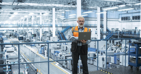 Experienced Male Engineer Standing on a Platform, Wearing a High-Visibility Vest, Using Laptop Computer and Overlooking Production at a Modern Automated Electronics Manufacture with Robotic Arms
