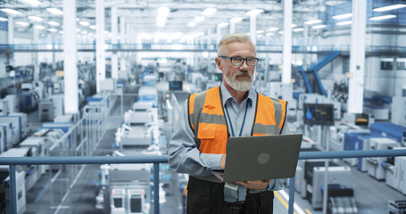Experienced Male Engineer Standing on a Platform, Wearing a High-Visibility Vest, Using Laptop Computer and Overlooking Production at a Modern Automated Electronics Manufacture with Robotic Arms - 792699657