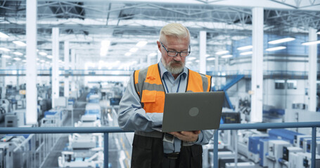 Senior Engineer Standing on a Platform, Using Laptop Computer at an Electronics Factory. Machines are Undergoing Maintenance, Specialist Monitoring the Updates Through Online Software