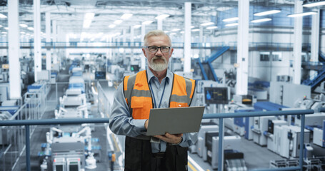 Portrait of a Handsome Middle Aged Engineer Wearing Glasses, Using Laptop Computer and Looking at Camera. Technician in High-VIsibility Vest Working in a Modern Technology Factory