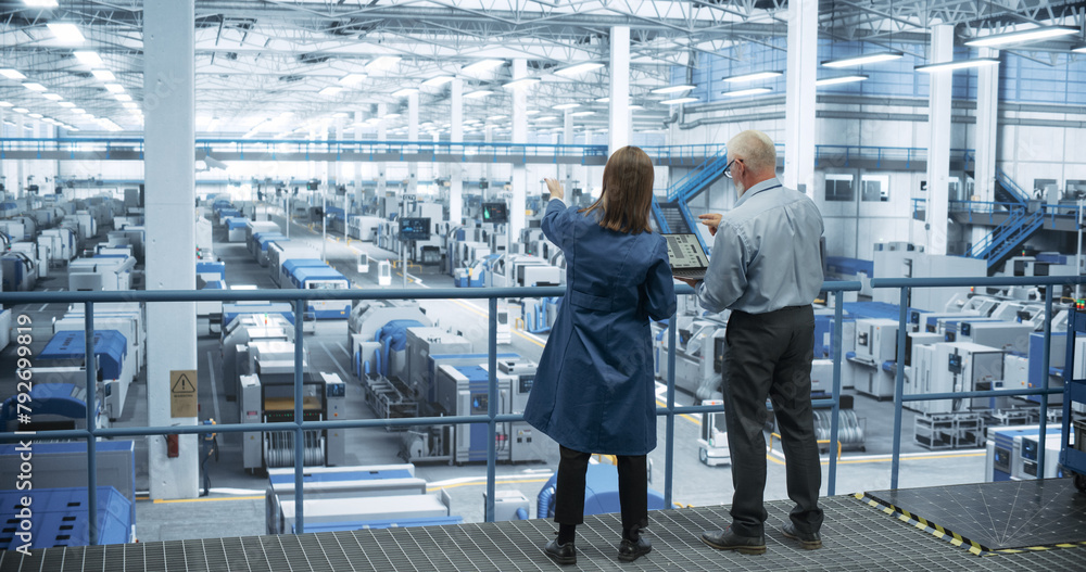 Poster male and female engineers standing with their back to camera, using laptop computer and talking in a