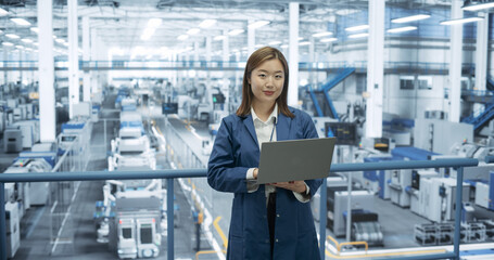 Portrait of a Young Asian Technician Using Laptop Computer and Looking at Camera. Industrial Female Specialist Wearing a Blue Coat, Working in a Modern Electronics Factory