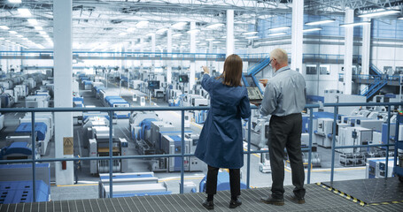 Male and Female Engineers Standing with Their Back to Camera, Using Laptop Computer and Talking in a Factory Facility with Equipment Producing Modern Electronic Components for Different Industries - 792699819