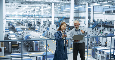 Multiethnic Male and Female Colleagues Standing in a Manufacturing Complex, Using Laptop Computer and Discussing Production on Modern Automated Electronic Devices and Gadgets