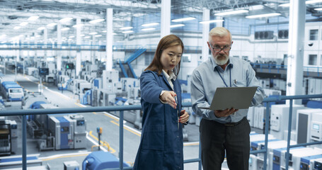 Middle Aged Engineer and Young Japanese Technician Having a Conversation and Using Laptop Computer. Specialists Monitoring, Analyzing Software Reports at a Modern Electronics Factory