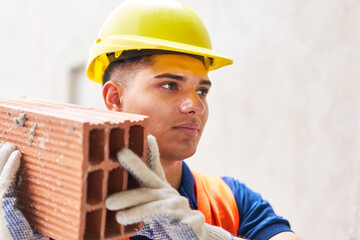 Confident young male bricklayer wearing safety hardhat carrying brick at construction site