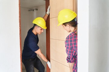 Young male and female construction workers wearing safety workwear installing door at incomplete house - obrazy, fototapety, plakaty