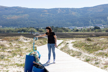 A conscious female helps clean the beach by picking up a blue piece of trash on a sunny day,...