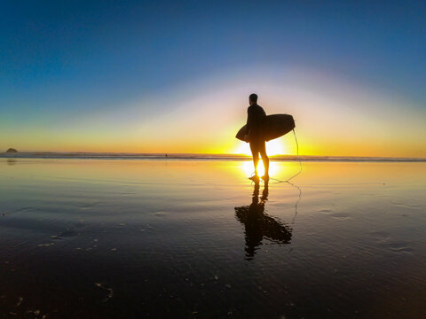 Silhouette of a surfer standing on the beach at sunset in New Zealand