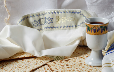 White talit with blue embroidery folded next to pieces of matzah and Kiddush cup filled with wine. Matzah pieces are flat and have a perforated pattern. The Kiddush cup, tallit. Religious celebration