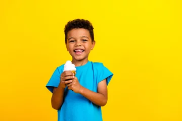 Foto op Plexiglas Portrait of pleasant little schoolboy with afro hair wear blue t-shirt holding ice cream in hands isolated on vivid yellow background © deagreez