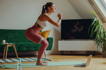 Sporty woman squats on mat in front of laptop in living room. Home workout concept
