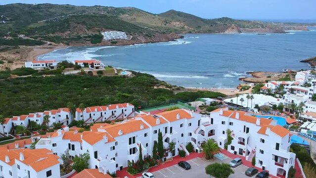 Aerial views of traditional white apartments hotel with red tiled roofs, swimming pool and tennis courts, with a beautiful sea cove in the background in Minorca, Spain