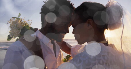 Image of light spots over happy african american bride and groom embracing on beach at wedding - obrazy, fototapety, plakaty