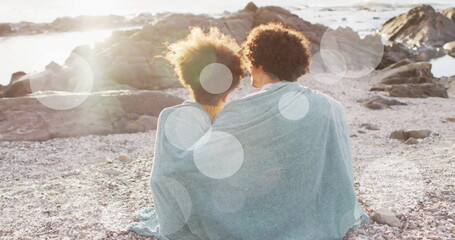 Naklejka premium Image of light spots over biracial couple covered in blanket embracing on beach