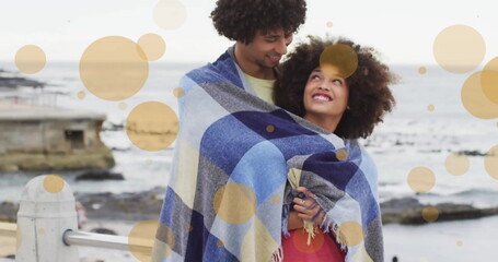 Fototapeta premium Image of light spots over biracial couple covered in blanket embracing on beach