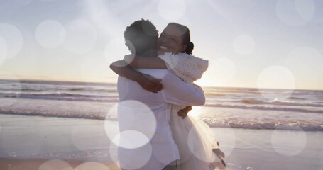 Obraz premium Image of light spots over happy african american bride and groom embracing on beach at wedding