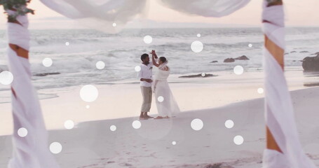 Fototapeta premium Image of white spots over african american bride and groom dancing on beach at wedding