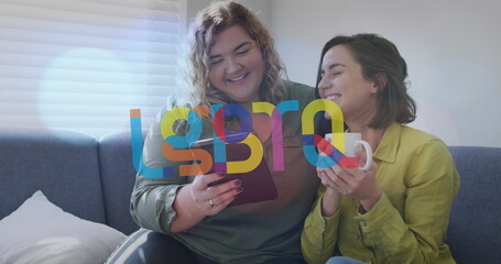 Image of lgbtq over happy caucasian lesbian couple using tablet and sitting on sofa at home