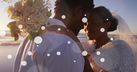 Image of white spots over happy african american bride and groom embracing on beach at wedding