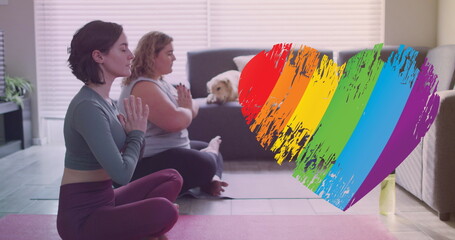 Image of rainbow heart over caucasian lesbian couple practicing yoga at home - obrazy, fototapety, plakaty