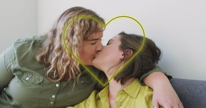 Image of neon heart over happy caucasian lesbian couple kissing at home
