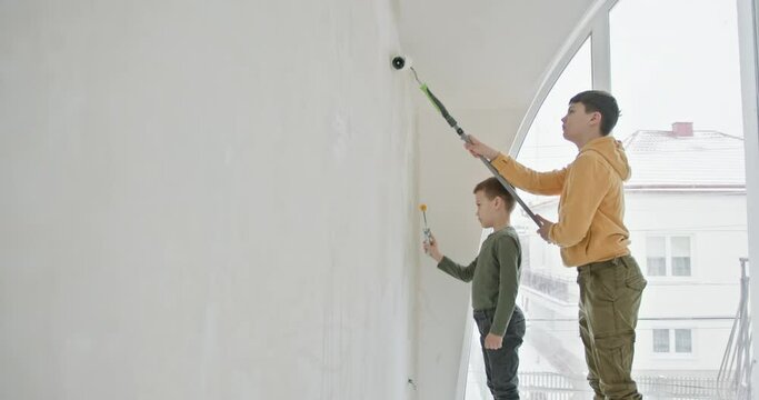 Two children are renovating their house by painting a large wall. They stand near a window, adding a feeling of lightness and space, apply paint with rollers. The moment of joint repair of the house