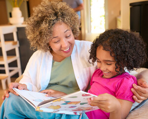 Grandmother Looking After Granddaughter At Home Sitting On Sofa Reading A Story Together
