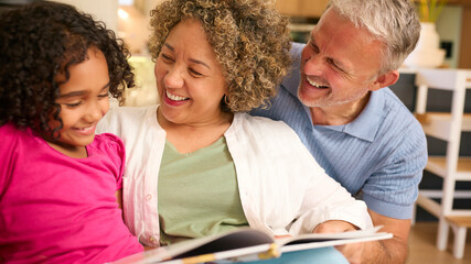 Multi Racial Grandparents Looking After Granddaughter At Home Reading A Story Together - obrazy, fototapety, plakaty