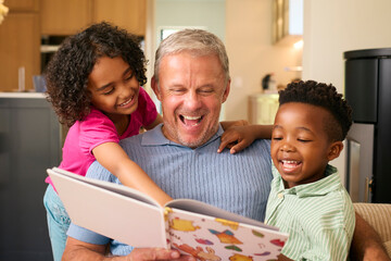 Multi Racial Family With Grandfather Looking After Grandchildren At Home Reading A Story Together - obrazy, fototapety, plakaty