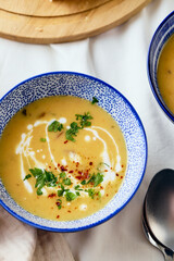 Two bowls of creamy potato soup, spoons, napkin and wooden cutting board with a loaf of bread and cut pieces. The surface of the soup is decorated with cream, croutons, parsley and ground red pepper.