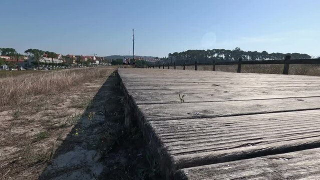 three people walk in the distance on the wooden planks of the overpass with old wooden railing on sandy soil in the direction of the city, shot from low turning right slow Pontevedra, Galicia, Spain