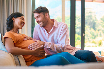 Loving Multi-Racial Couple With Pregnant Woman On Sofa And Man Touching Woman's Stomach At Home