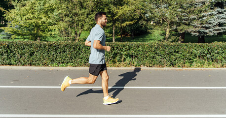 A runner in stride, his shadow cast on the park's path, is surrounded by verdant shrubs under the...