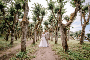 Valmiera, Latvia - August 10, 2023 - Newlyweds stand on a tree-lined path, the groom holding the...