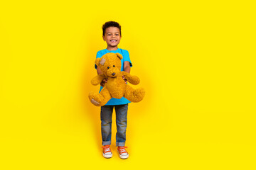Full length photo of positive child with curly hair dressed blue t-shirt denim jeans hold teddy bear in arms isolated on yellow background