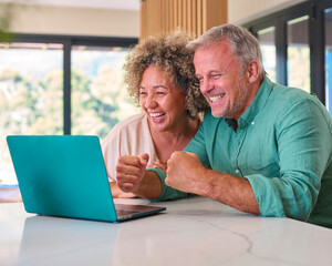 Multi-Racial Mature Couple At Home Celebrating Good News Looking At Laptop