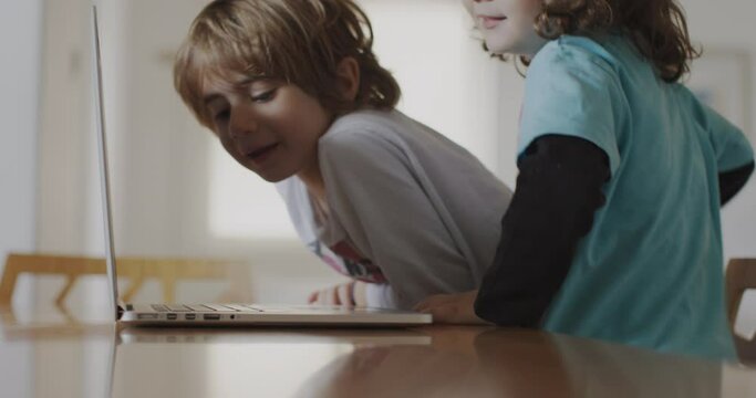 Two small boys are sitting at a table in their living room, engrossed on a laptop screen.