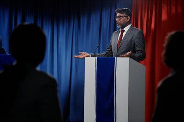 Confident mature male representative of government speaking to foreign colleague while standing by platform at conference