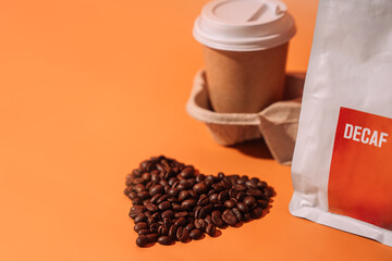 Coffee beans in the shape of a heart on an orange background next to packaging with text decaf and a paper cup in a tray.