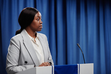 Young female politician looking at opponent during political debating or at conference while standing by platform with microphone