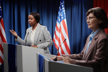 Confident young female delegate standing by platform against American flag in conference hall and answering question of press