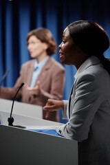 Focus on young confident female politician speaking in microphone while standing by platform in conference hall and debating with public